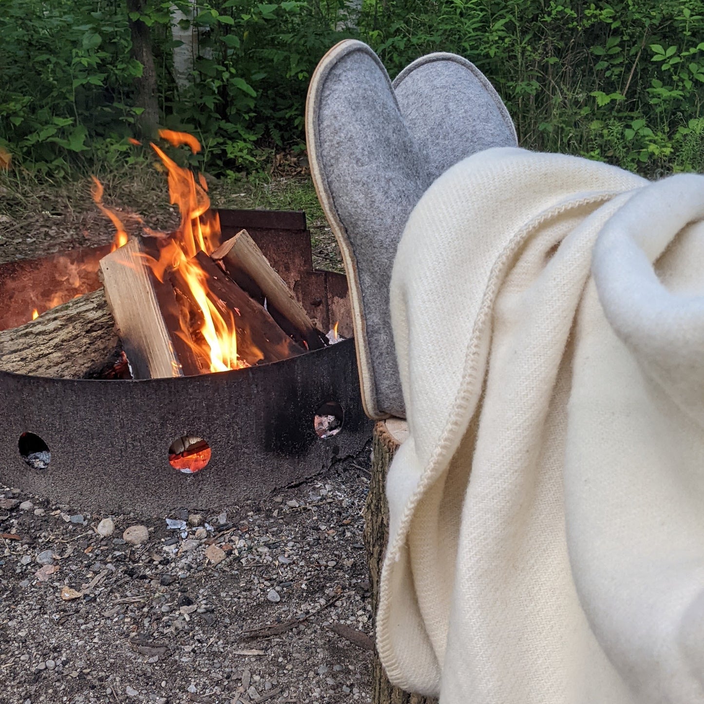 Person sitting by a campfire in a wooded area, wearing slippers and a blanket.